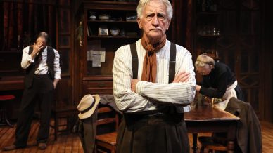 An older man stands with arms crossed and a serious expression in the centre of a rustic kitchen. Two other people are behind him; one covers his face, the other leans over a table. The setting is warm and wooden.