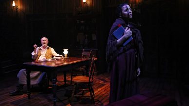 A dimly lit stage shows an older man sitting at a desk with an open book and small lamp, looking at a woman standing nearby holding a closed book, dressed in dark, old-fashioned clothes.
