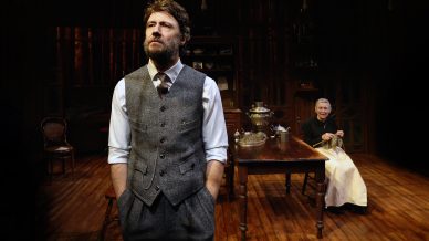 A man in a grey waistcoat and tie stands at the front of a wooden stage, looking serious. Behind him, an older woman sits at a table, knitting and smiling, with shelves and chairs in the background.