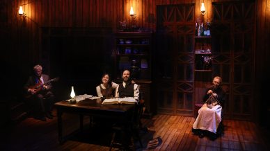 Four people sit on a dimly lit wooden stage set resembling a rustic room; a man plays guitar on the left, a couple sits at a table with books at centre, and an older woman knits on the right near shelves and oil lamps.