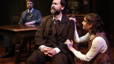 Three people in period clothing are on a stage set with wooden furniture. A bearded man sits at the centre, looking serious, while a woman kneels beside him, touching his shoulder. Another man sits at a table in the background.