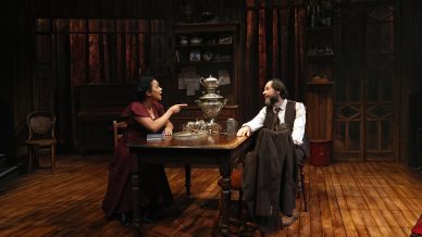 Two actors in period costumes sit at a wooden table on a dimly lit stage set, featuring vintage furniture and a samovar. The woman points at the man as they engage in conversation. The room has wooden floors and shelves in the background.
