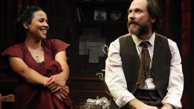 A woman in a burgundy dress smiles at a bearded man in a waistcoat and tie. They sit side by side onstage against a dark, antique backdrop, suggesting a scene from a theatrical play.