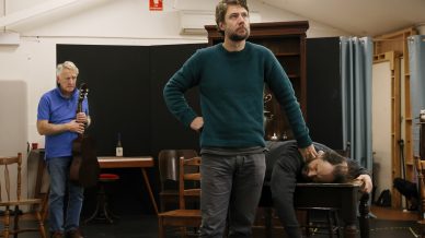 Three men in a rehearsal room: one stands with a guitar in the background, one stands centre stage looking ahead, and another lies face-down on a table as if unconscious. Chairs and a bottle are scattered around.