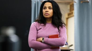 A woman with long dark hair wearing a purple jumper stands indoors with her arms crossed, holding a red book, looking thoughtful. Blue curtains and a black panel are in the background.