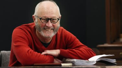 An older man with glasses and grey sideburns, wearing a red jumper, sits at a table smiling, with his arms crossed over an open notebook and some papers.