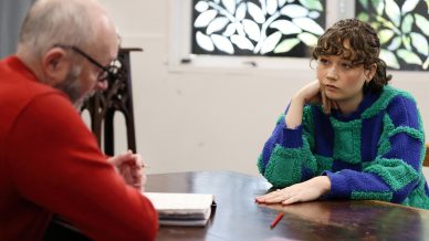 A young woman in a blue and green jumper sits at a wooden table, resting her head on her hand, looking thoughtful while listening to an older man with glasses who is writing in a notebook.