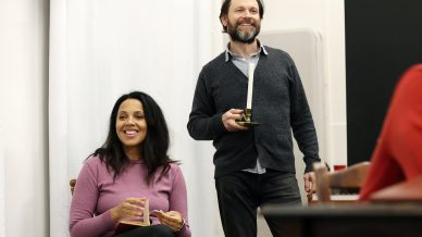 A woman in a pink jumper sits smiling with a book and mug, while a bearded man in a grey cardigan stands nearby holding a candlestick, both appearing cheerful in a bright room with white curtains.