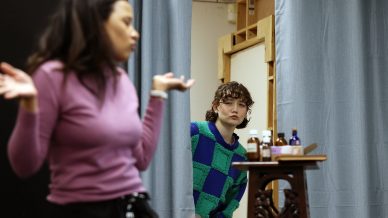 A person in a chequered green and blue jumper stands in a doorway, looking at another person in a pink top who is shrugging with hands raised. Various bottles sit on a wooden table in the foreground.