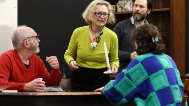 Four adults are gathered around a wooden table. A woman in a green jumper stands holding a candle, appearing concerned. Two men and another woman sit nearby, engaged in conversation. The setting looks like a dramatic scene or rehearsal.