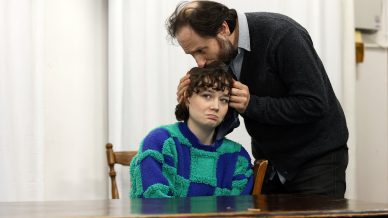A man gently kisses the head of a young person with short curly hair, who is sitting at a table and looking sad while wearing a blue and green checked jumper. The background is plain with white curtains.