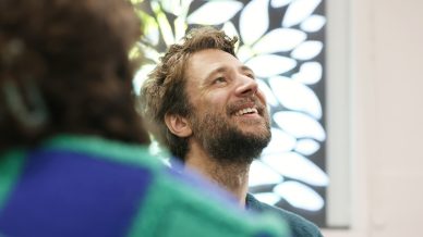 A bearded man smiles and looks upwards, sitting indoors with a leafy window pattern in the background. The blurred shoulder of another person in a green and blue jumper is visible in the foreground.