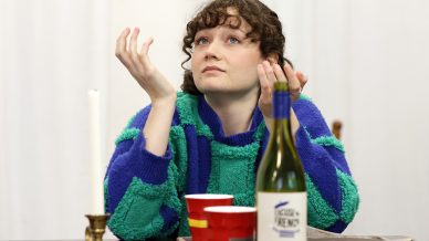 A person with curly hair in a blue and green jumper sits at a table with a wine bottle, red cups, a candle, and a tray, looking up with hands raised in an expressive gesture.