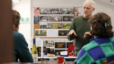 An older man in a green jumper speaks to two people sitting at a table in a classroom or workshop. The background shows photos and notes pinned to a board, and there are bottles and papers on the table.