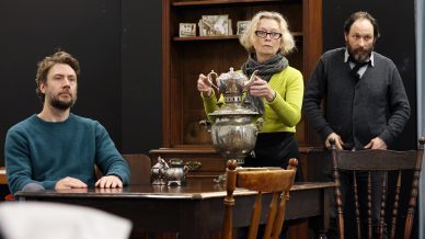 Three adults in a room: one man sits at a wooden table, a woman in glasses stands holding a large silver teapot on a samovar, and another man stands behind them. Shelves and framed photos are in the background.