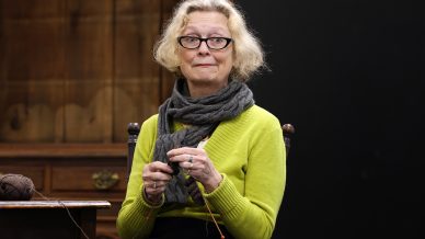 An older woman with fair hair and glasses wears a green jumper and dark scarf, sitting and knitting with grey wool. She is indoors, in front of a wooden cupboard and a dark background.