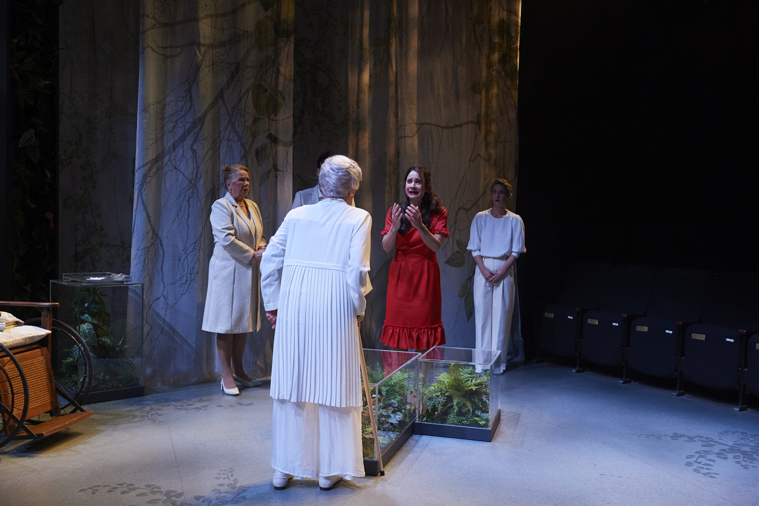 Four women stand on a stage with forest-themed curtains and plants in glass cases. One woman in a red dress gestures expressively whilst the others, dressed in light-coloured clothing, listen. The scene is softly lit.