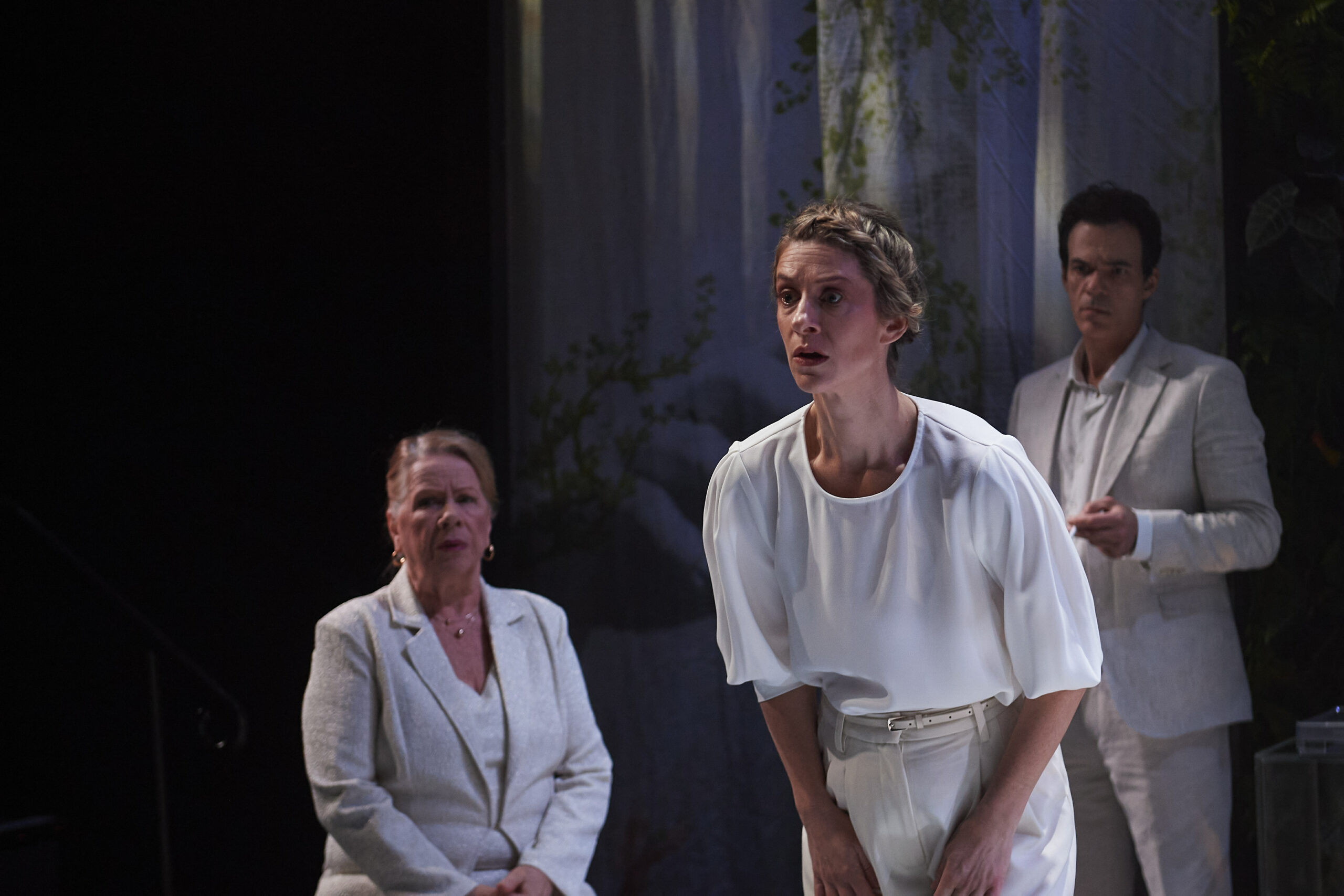 A woman in white leans forward, looking intently ahead, while an older woman and a man, also dressed in white, watch her. They appear to be on stage with a forest backdrop, suggesting a dramatic scene in a play.