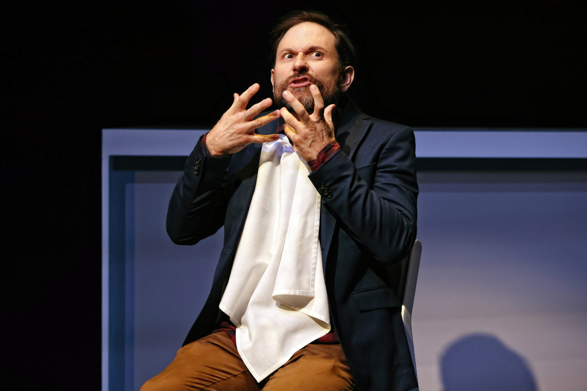 A man with a serviette tucked into his shirt sits on a chair, gesturing dramatically with both hands near his face, eyes wide and mouth slightly open, as if expressing surprise or excitement.