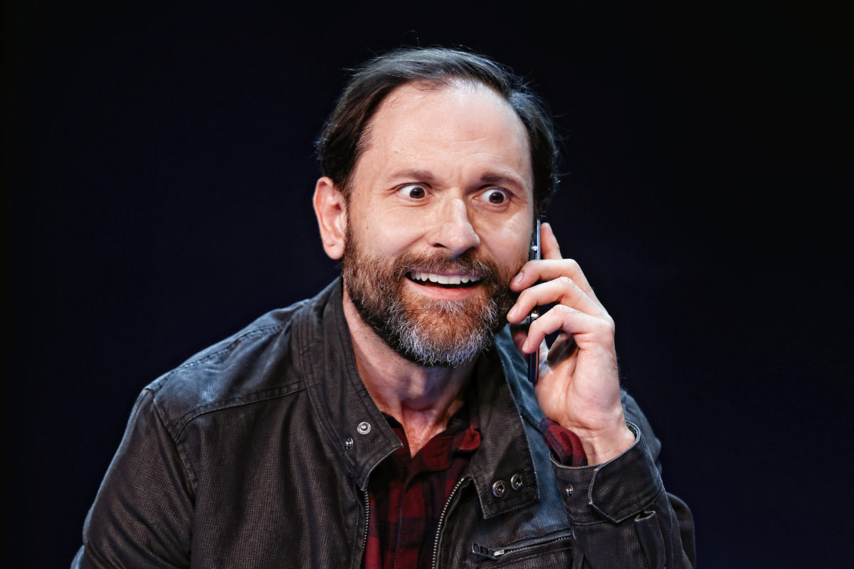 A bearded man with a slight smile and wide eyes talks on a mobile phone. He wears a dark jacket over a red checked shirt and is set against a dark background.